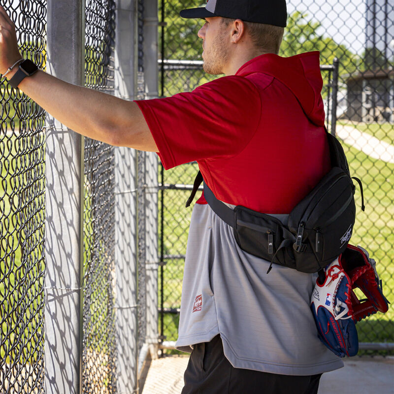 A player standing in a dugout with a black Rawlings sling bag on his back - SKU: RSLINGBG